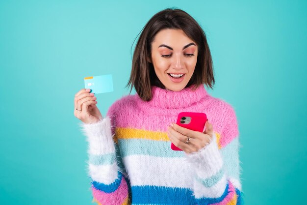 Young woman in a bright multicolored sweater on blue holds a credit card