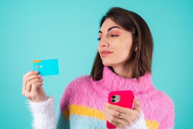 Young woman in a bright multicolored sweater on blue holds a credit card