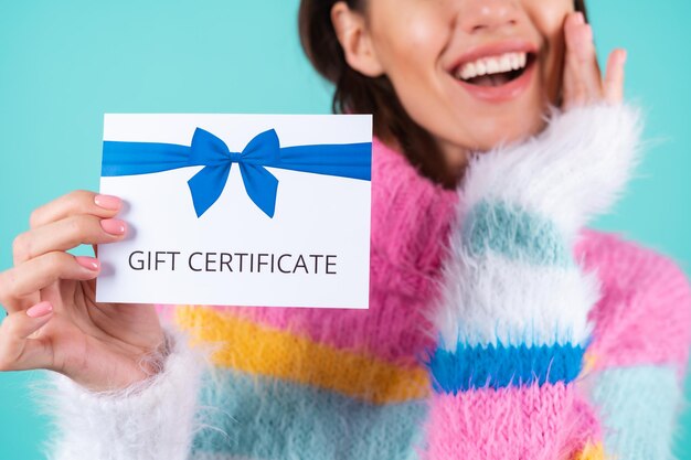 Young woman in a bright multicolored sweater on blue holds a gift certificate, smiles enthusiastically