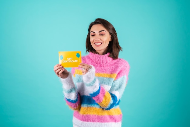 Young woman in a bright multicolored sweater on blue holds a gift certificate