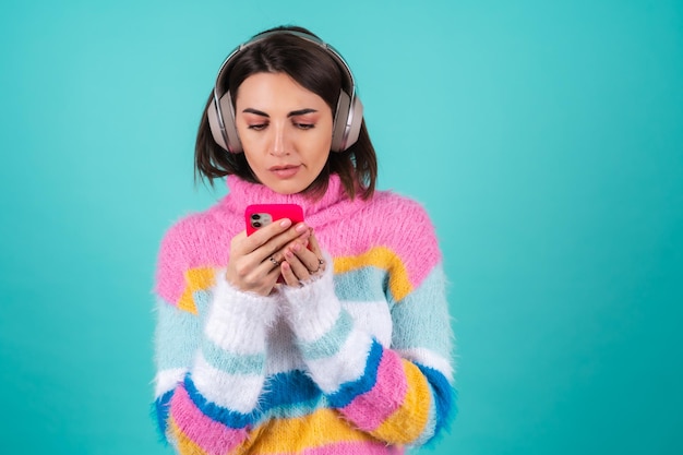 La giovane donna in un maglione multicolore luminoso sul blu in grandi cuffie di cancellazione del rumore tiene un telefono con uno sguardo annoiato dispiaciuto