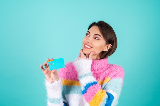 A young woman in a bright multicolored sweater on blue shows a credit card, looks thoughtfully