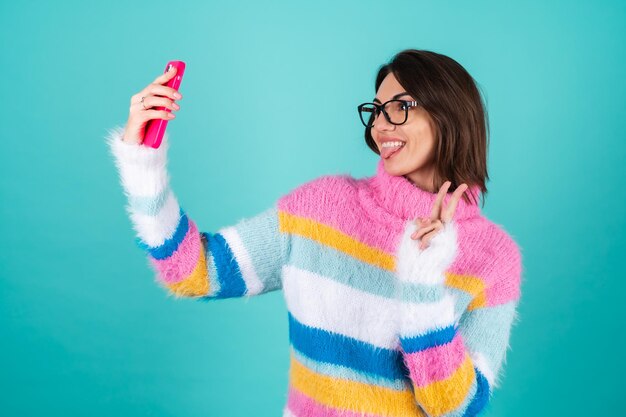 A young woman in a bright multicolored sweater on blue with glasses, holds a phone, takes a selfie