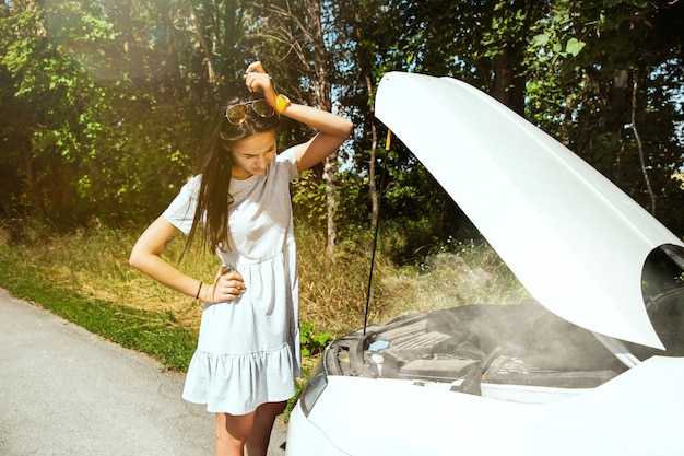 若い女性は休憩の途中で車を壊した。彼女は自分で壊れたものを直そうとしている、またはヒッチハイクする必要があります。緊張する。週末、道路のトラブル、休暇。