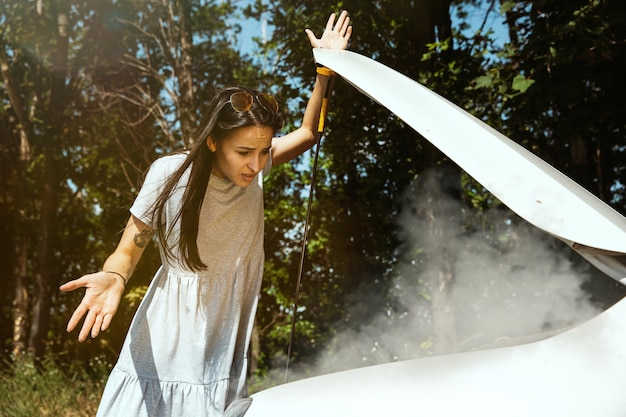 若い女性は休憩の途中で車を壊した。彼女は自分で壊れたものを直そうとしている、またはヒッチハイクする必要があります。緊張する。週末、道路のトラブル、休暇。