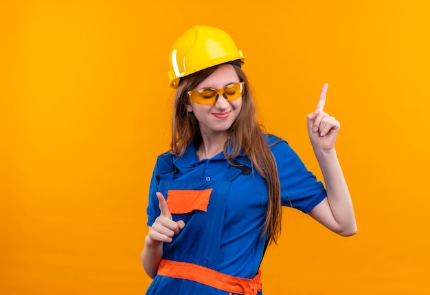 オレンジ色の壁の上に立って元気に人差し指を指して笑顔を楽しんでいる建設制服と安全ヘルメットの若い女性ビルダー労働者