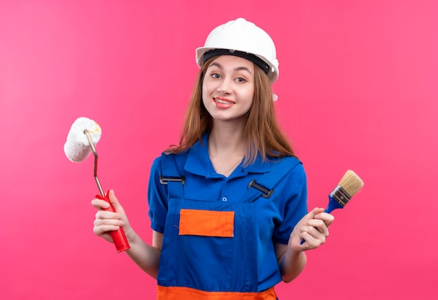 Operaio del costruttore della giovane donna in uniforme da costruzione e casco di sicurezza che tiene pennello e rullo di vernice sorridente in piedi amichevole sopra la parete rosa