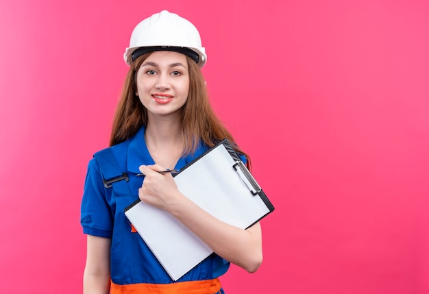 Operaio del costruttore della giovane donna nella lavagna per appunti della tenuta del casco di sicurezza e dell'uniforme della costruzione che sembra sicura che si leva in piedi sopra la parete rosa