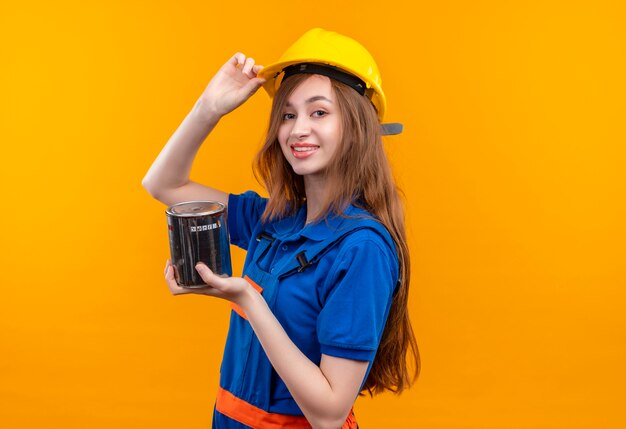 Young woman builder worker in construction uniform and safety helmet holding paint can looking confident touching her helmet standing over orange wall