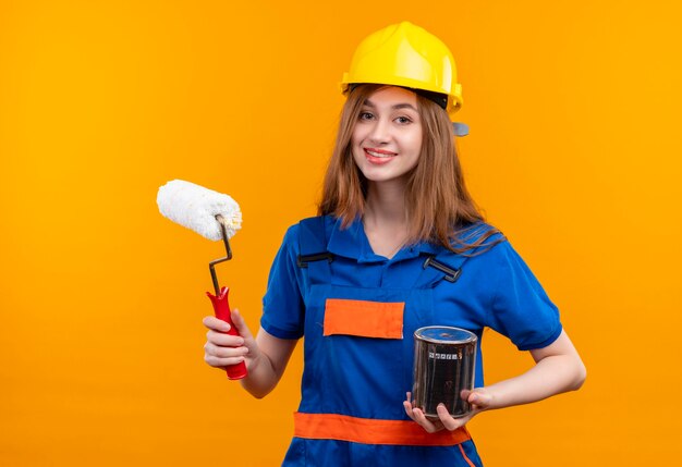 Молодая женщина-строитель в строительной форме и защитном шлеме, держащая банку с краской и улыбающийся ролик
