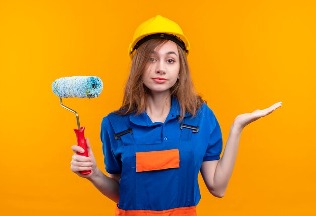 Young woman builder worker in construction uniform and safety helmet holding paint roller looking confused shrugging standing over orange wall