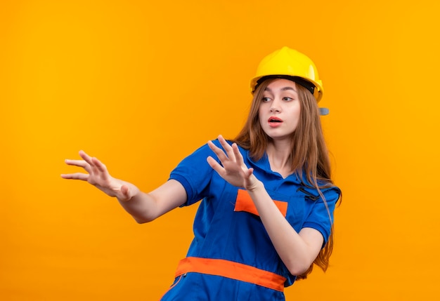 Young woman builder worker in construction uniform and safety helmet holding palms up making defense gesture standing over orange wall