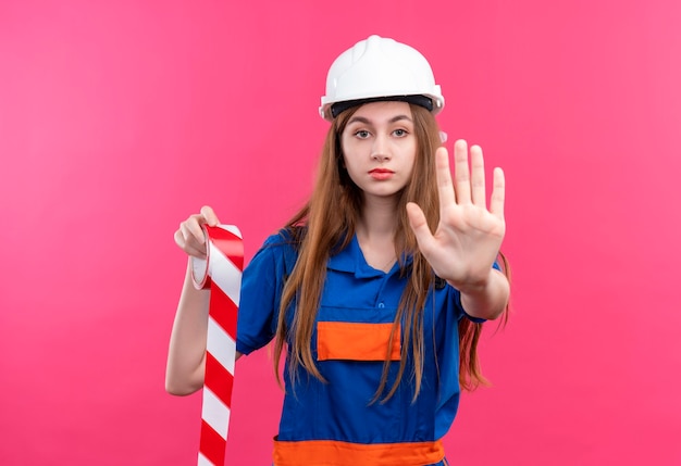 Young woman builder worker in construction uniform and safety helmet holding scotch tape making stop sign with hand standing over pink wall