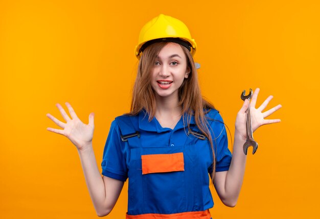 Young woman builder worker in construction uniform and safety helmet holding wrench raising hands in surrender smiling standing over orange wall