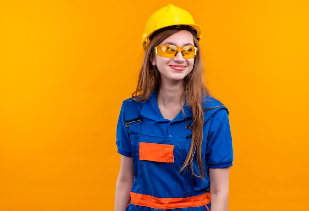 建設制服と安全ヘルメットの若い女性ビルダー労働者は元気に立って笑顔を脇に見て