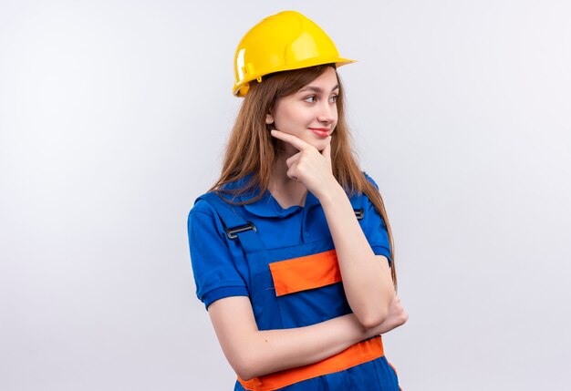 Young woman builder worker in construction uniform and safety helmet looking aside  standing with hand on chin with pensive expression thinking over white wall