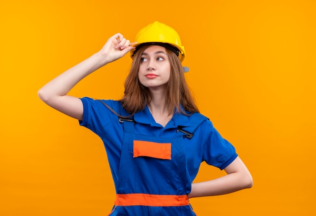 Young woman builder worker in construction uniform and safety helmet looking aside with confident look touching her helmet standing