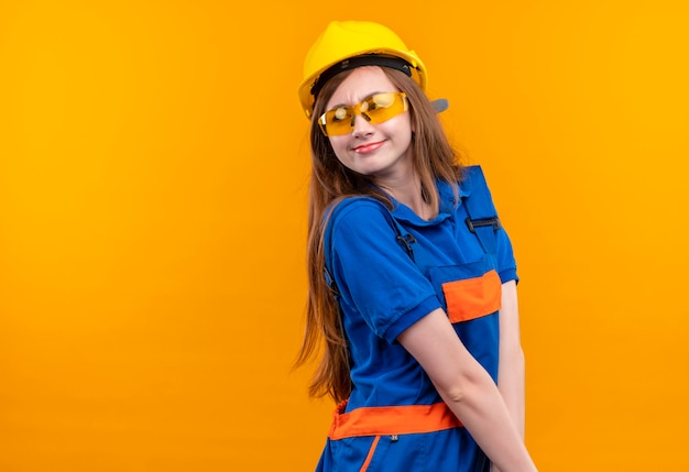 Young woman builder worker in construction uniform and safety helmet looking aside with shy smile on face standing