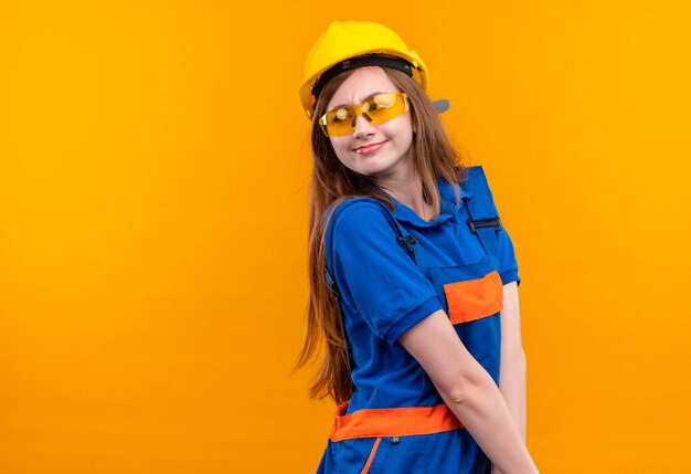 Молодая женщина-строитель в строительной форме и защитном шлеме смотрит в сторону с застенчивой улыбкой на лице