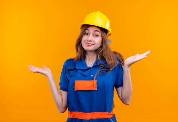 Молодая женщина-строитель в строительной форме и защитном шлеме выглядит смущенной, улыбаясь, пожимая плечами, не имея ответа стоя