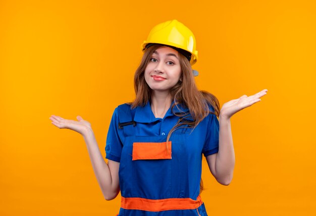 Young woman builder worker in construction uniform and safety helmet looking confused smiling shrugging shoulders, having no answer standing