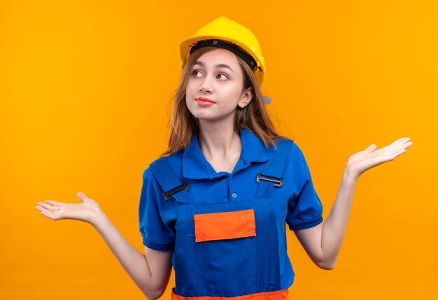 Young woman builder worker in construction uniform and safety helmet looking confused smiling shrugging shoulders, having no answer standing