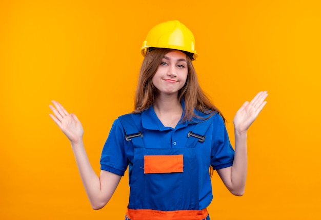 Young woman builder worker in construction uniform and safety helmet looking confused spreading arms to the sides standing