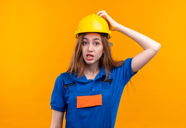 Young woman builder worker in construction uniform and safety helmet looking confused touching helmet for mistake standing
