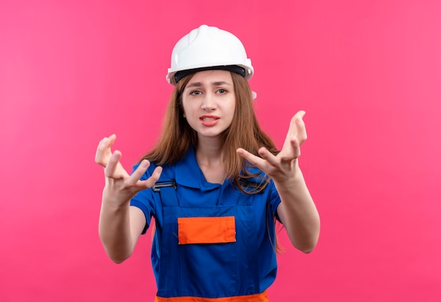Young woman builder worker in construction uniform and safety helmet looking disappointed raising hands standing over pink wall