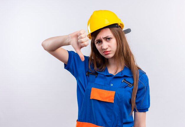 白い壁の上に立って親指を下に見せて不機嫌そうに見える建設制服と安全ヘルメットの若い女性ビルダー労働者
