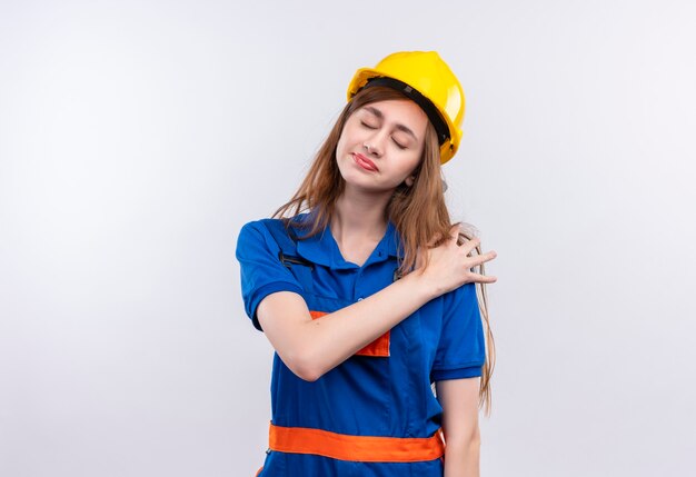 건설 유니폼 및 안전 헬멧에 젊은 여자 작성기 작업자 흰 벽 위에 서있는 고통을 느끼고 피곤 감동 어깨를 찾고