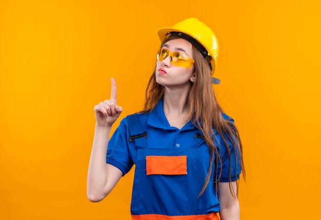 Young woman builder worker in construction uniform and safety helmet looking up intrigued pointing with index finger to something standing