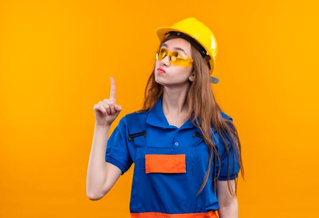 Young woman builder worker in construction uniform and safety helmet looking up intrigued pointing with index finger to something standing