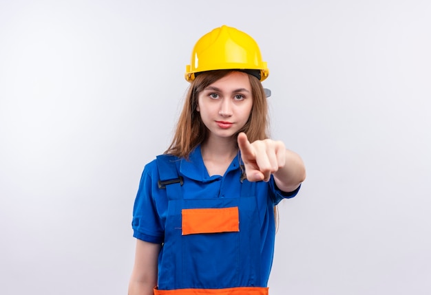 Young woman builder worker in construction uniform and safety helmet looking with serious face pointing with index finger