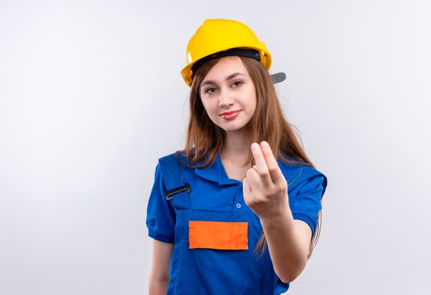 Young woman builder worker in construction uniform and safety helmet making come in gesture with hand looking confident standing over white wall