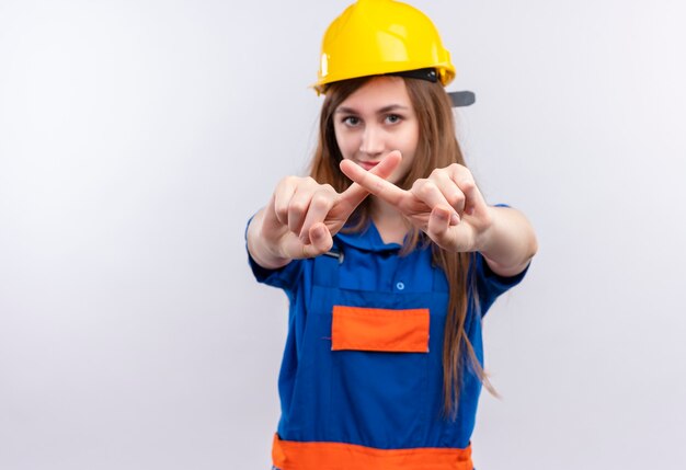 Young woman builder worker in construction uniform and safety helmet making defense gesture crossing index fingers  standing over white wall