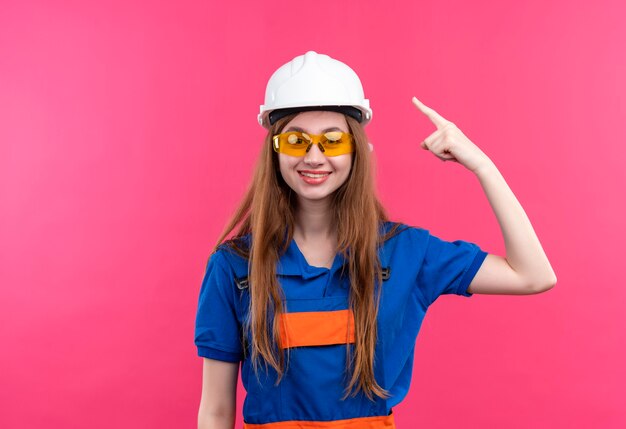 Young woman builder worker in construction uniform and safety helmet pointing index finger to head