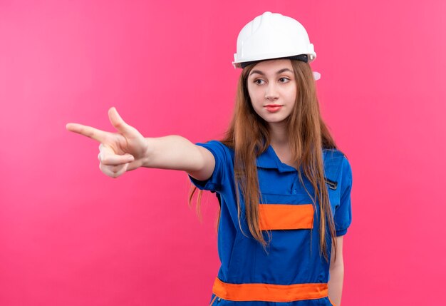 Young woman builder worker in construction uniform and safety helmet pointing to something with serious face standing over pink wall
