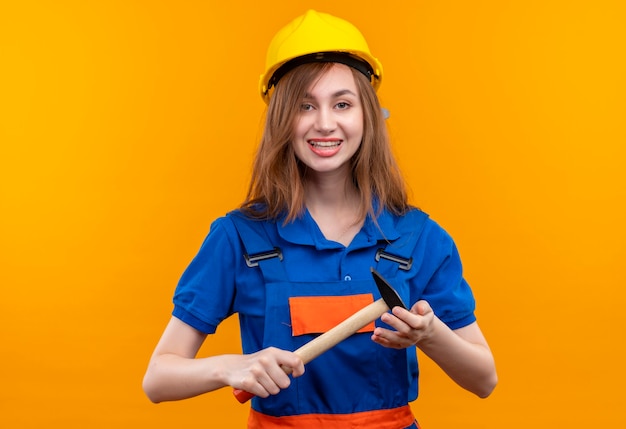 Young woman builder worker in construction uniform and safety helmet positive and happy smiling holding hammer standing over orange wall