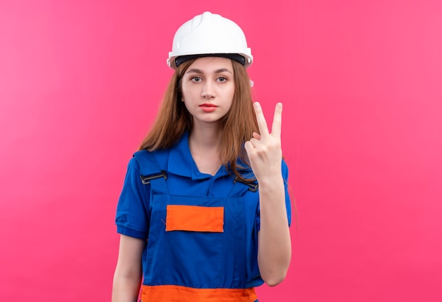建設制服と安全ヘルメットの若い女性ビルダー労働者が2番目の指で上向きに見せて