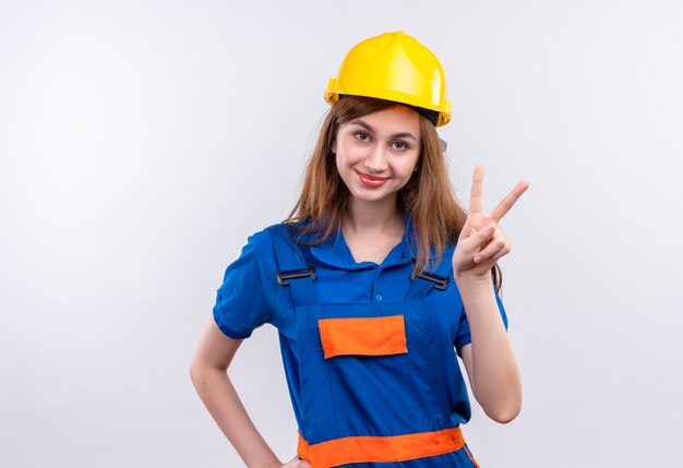 建設制服と安全ヘルメットの若い女性ビルダー労働者は、白い壁の上に友好的に立って笑顔の勝利のサインを示しています