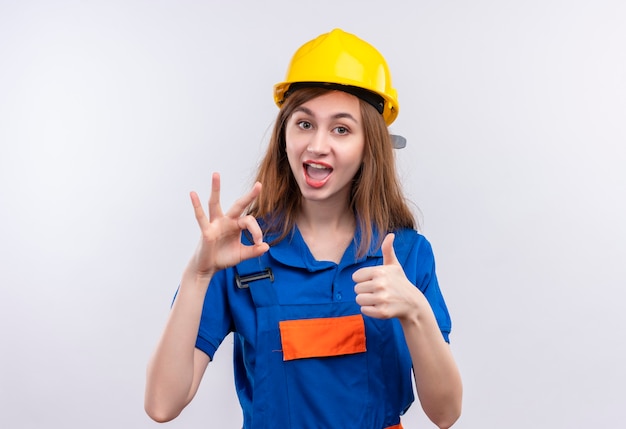 건설 유니폼과 안전 헬멧에 젊은 여자 작성기 노동자는 광범위하게 엄지 손가락을 보여주는 웃 고 흰 벽 위에 서 확인 서명