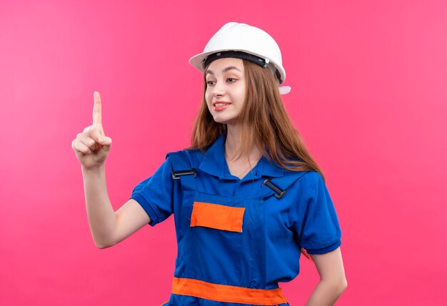 건설 유니폼 및 안전 헬멧에 젊은 여자 작성기 노동자 핑크 벽 위에 서 자신감 보여주는 검지 손가락 미소