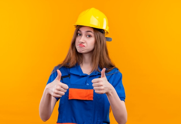 Молодая женщина-строитель в строительной форме и защитном шлеме, уверенно улыбаясь, показывает большие пальцы вверх обеими руками, стоя над оранжевой стеной