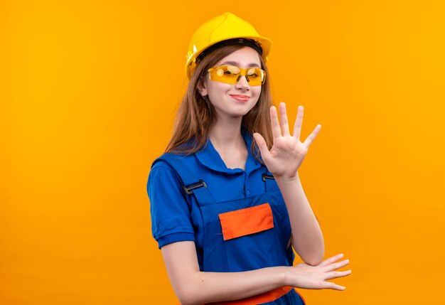 オレンジ色の壁の上に立っている手で自信を持って手を振って笑顔の建設制服と安全ヘルメットの若い女性ビルダー労働者