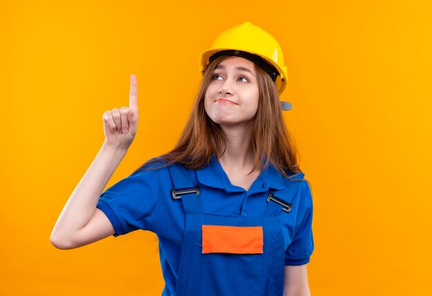 オレンジ色の壁の上に立って人差し指を笑顔で建設制服と安全ヘルメットの若い女性ビルダー労働者