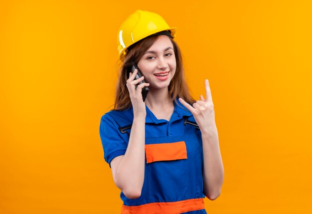 オレンジ色の壁の上に立っている指で岩のシンボルをやって、携帯電話で話している間笑顔の建設制服と安全ヘルメットの若い女性ビルダー労働者