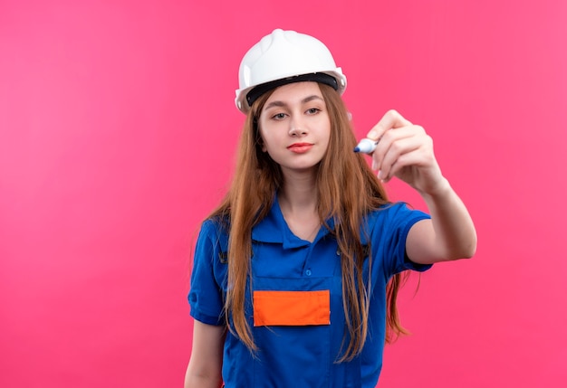 Young woman builder worker in construction uniform and safety helmet trying to write something in air with pen standing over pink wall