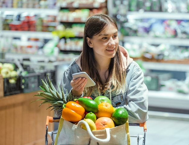 A young woman buys groceries in a supermarket with a phone in her hands.