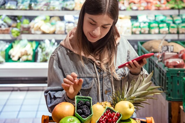 Молодая женщина покупает продукты в супермаркете с телефоном в руках.